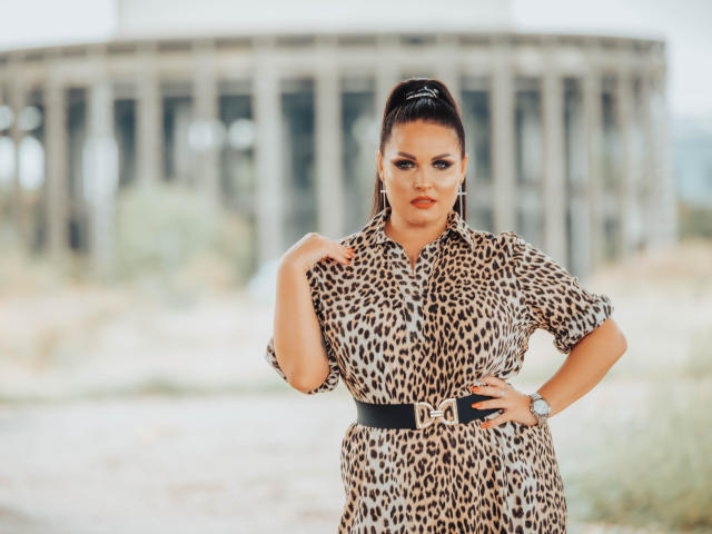 GeorgiaLerox trägt ein Kleid mit Leopardenmuster, einen schwarzen Gürtel und eine Uhr, steht mit einer Hand am Hüfte, hat dunkles Haar zu einem hohen Pferdeschwanz gebunden, wirkt selbstbewusst und hat einen ernsten Gesichtsausdruck..