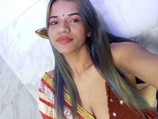 KairaDayamai with long, straight, dark green hair, wearing a red dress with gold and white patterns, showing her tits in a low-cut dress, smiling, white tile wall background.