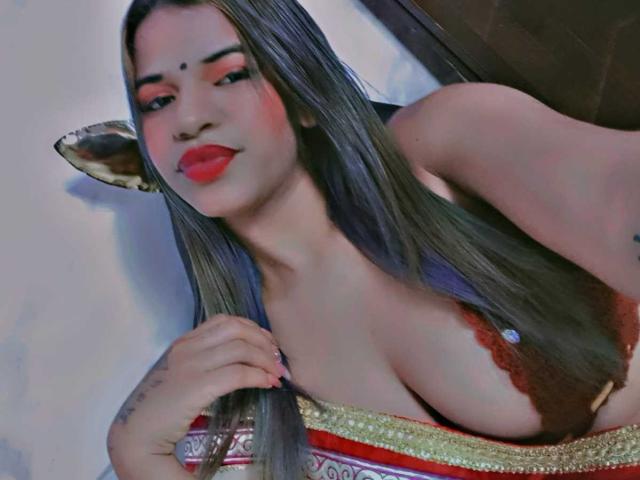 KairaDayamai with long grayish-brown hair, red lipstick, and a brown lace bra showing her big tits, holding her hair near her cleavage, wearing a colorful patterned top.