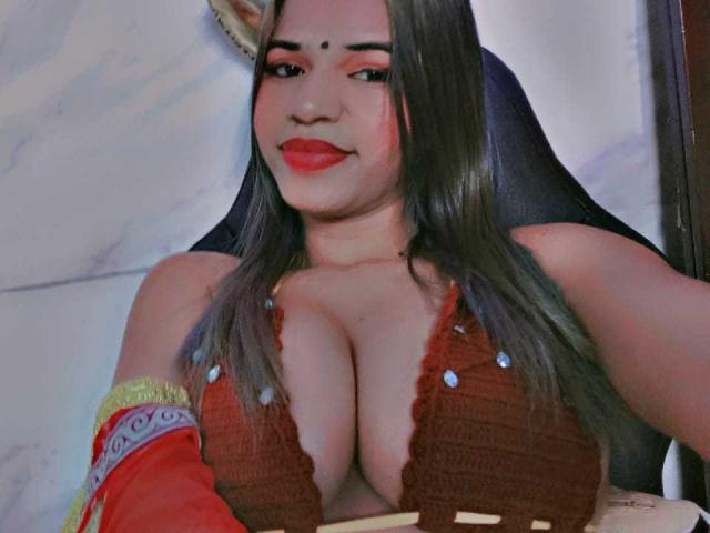 KairaDayamai has big, round tits in a red, crocheted top, with long brown hair, red lipstick, and a small black dot on her forehead. She's smiling slightly.