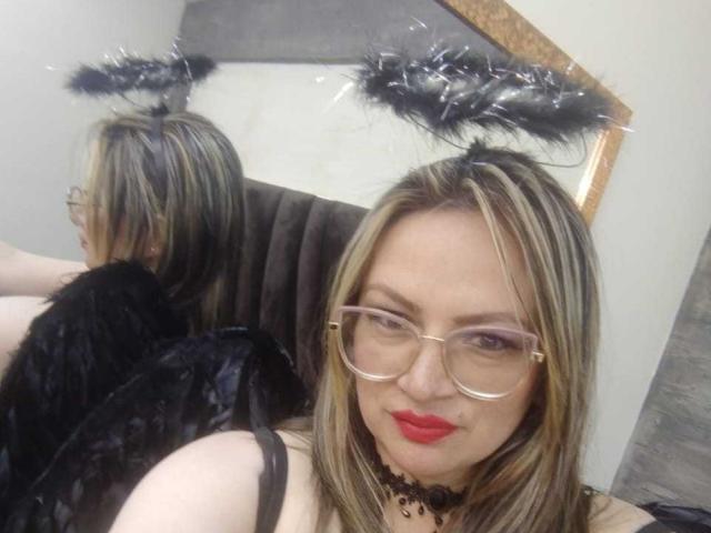 LadyLure with blonde hair, red lipstick, glasses, black angel wings, and black halo headpiece, showing her back and smiling at the camera, reflecting in a mirror.
