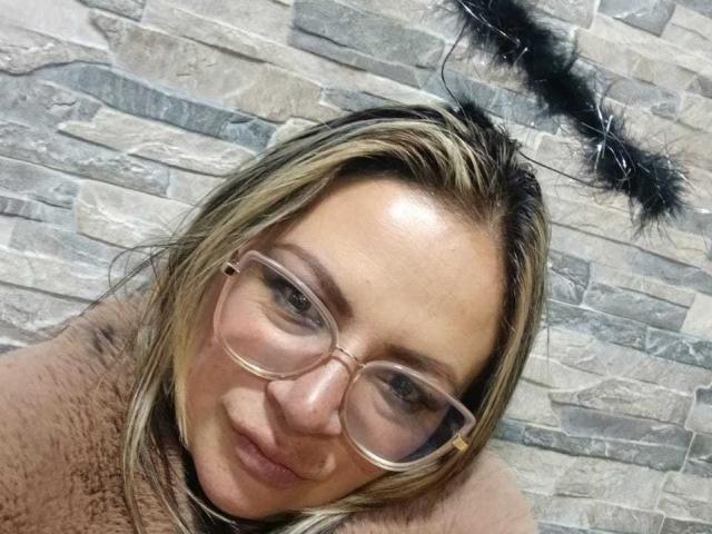LadyLure has blonde hair, glasses, and a slight smile with a fluffy pink top. Her eyes are focused, and a black feather is on her head.