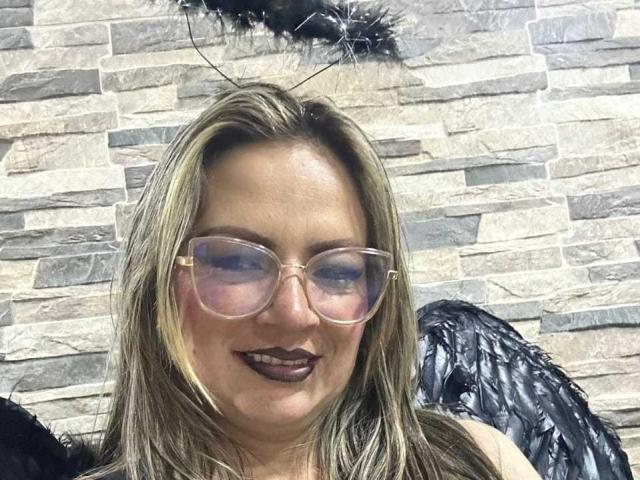 LadyLure with blonde hair, glasses, dark lipstick, and black wings, smiling at the camera, wearing a feathered headpiece. Her tits are not visible, only her face and wings.