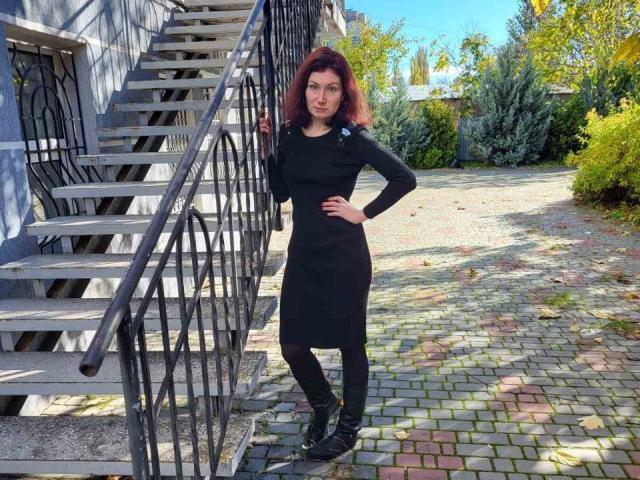AmiraGold stands on a cobbled path, leaning on a metal staircase. She's wearing a black dress, black leggings, and black boots, with red curly hair and a confident smile.