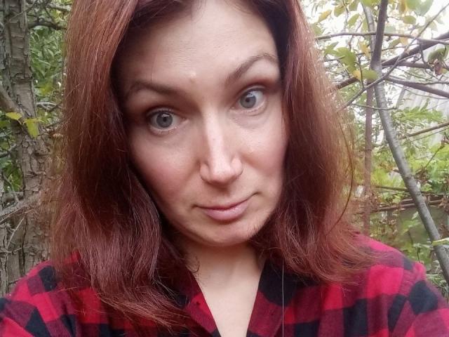 AmiraGold has red hair, green eyes, and a slight smile. She's wearing a red and black checkered shirt. Her nose has a small, visible blemish.