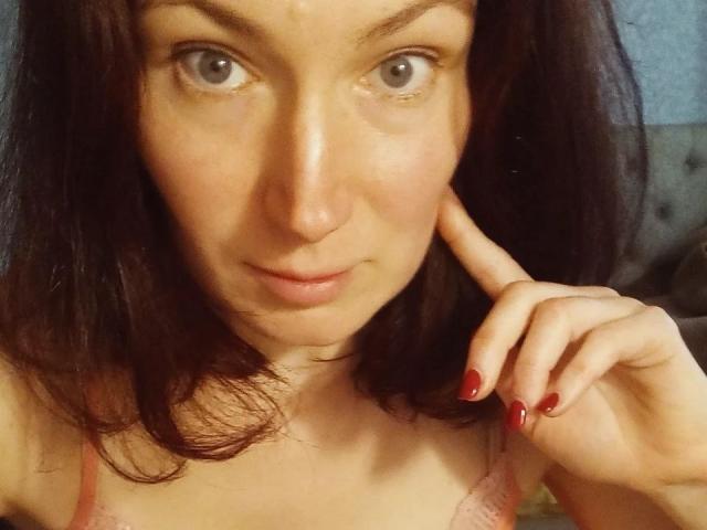 AmiraGold has gray eyes, fair skin, and red-painted nails. She's touching her chin with her right hand, showing off her tits in a pink lace bra.