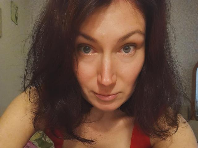 AmiraGold has dark red, slightly messy hair and blue eyes, with a subtle smile and visible nose, wearing a red top, showing her bare shoulders and chest.