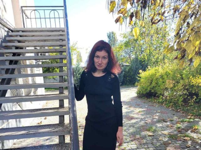 AmiraGold with red hair stands wearing a black dress, her hand on a metal staircase, cobblestone path, and greenery in the background. She's looking slightly to the side.