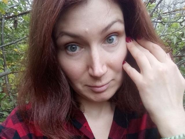AmiraGold has light green eyes, red hair, and is wearing a red and black plaid shirt. She's touching her face with red-painted nails. Her tits are small.