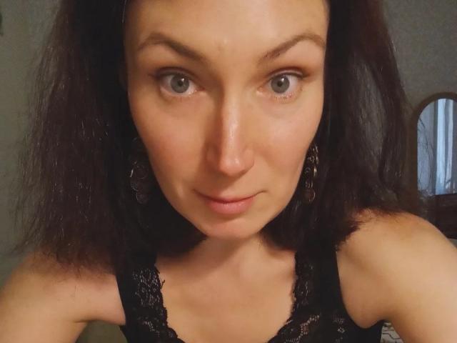 AmiraGold has gray eyes, dark brown shoulder-length hair, and is wearing black lace lingerie with matching earrings, showing off her small tits and slightly parted pink lips.