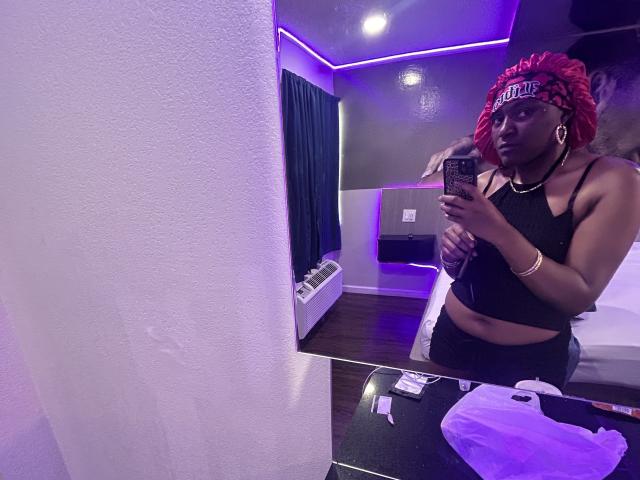 HoneyMakeMeCum with red braided hair, black crop top, and black underwear, holding phone, purple room, white wall, black table with bag, and phone charger in mirror selfie.