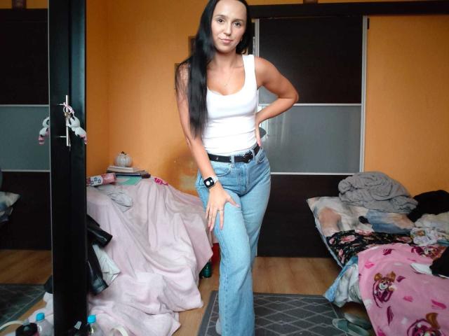 LuisaKiss stands in a cluttered bedroom. She has long black hair and is wearing a white tank top with a black belt and blue jeans. Her right hand rests on her hip, and her left hand is on her thigh. She has a black wristband on her left wrist. The room has orange walls, a messy bed with pink and black patterned covers, and a pile of clothes. A mirror is partially visible in the background. The floor is wooden.