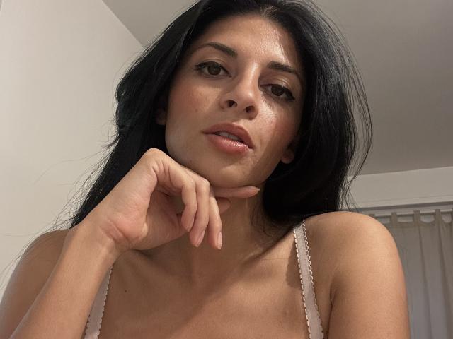 Rodalinda with long black hair, brown eyes, and light skin, wearing a white bra, has her chin resting on her hand, looking slightly to the side, with a subtle smile.