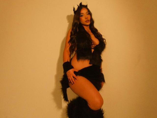 LucyCarson with long dark hair, black devil horns, and furry black outfit, showing her tits and ass, wearing black wrist cuffs, and furry leg cover, against a plain wall.
