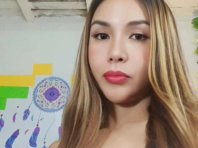 MissHotAyaLove has long blonde hair, pink lipstick, and brown eyes, staring at the camera with a neutral expression, her bare shoulders visible. Her tits are small, and she's looking cute.