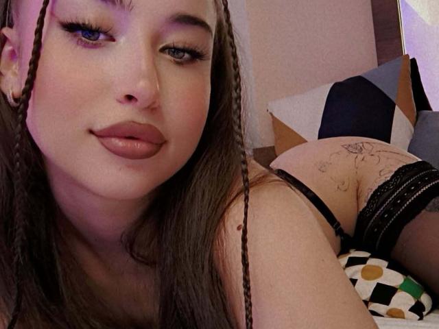 AdacGrey, com cabelo trançado, lábios cheios e lingerie preta, exibindo sua bunda tatuada em uma foto. Seus olhos azuis e piercing no nariz são visíveis. Seus seios estão parcialmente à mostra..