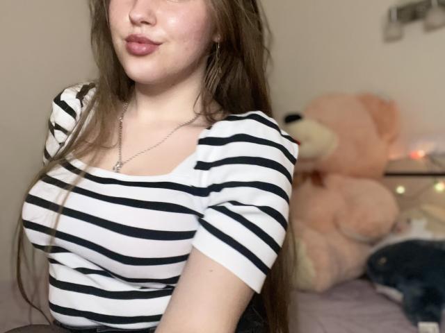 Dooris with long brown hair, wearing a striped white and black shirt, showing her tits, pouty lips, and a silver necklace, sitting in a bedroom with a large pink teddy.