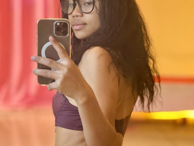 AubreyLoo takes a selfie, holding a smartphone with a circular ring on the back. She has long, wavy black hair and wears large, black-rimmed glasses. Her lips are slightly parted, and she has a light brown skin tone. She's wearing a purple sports bra, revealing a slim, athletic build. The background is blurred, featuring red and yellow colors. The image is a realistic, candid photograph.