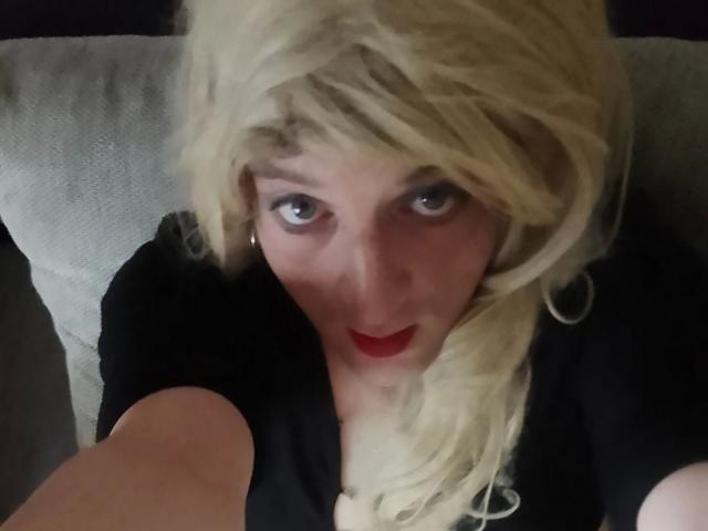 ZackaSissy20-hot hat blondes Haar, roten Lippenstift, ein schwarzes Oberteil und blickt mit blauen Augen nach oben. Sie hält einen Selfie-Stick und zeigt ihr Gesicht und ihren Oberkörper..