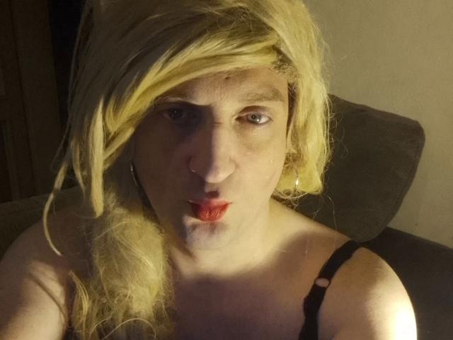 ZackaSissy20 – attraktiv mit blonder Perücke, Nasenring und rotem Lippenstift, mit leicht geöffnetem Mund, trägt ein schwarzes Top mit Trägern, in einem schwach beleuchteten Raum..