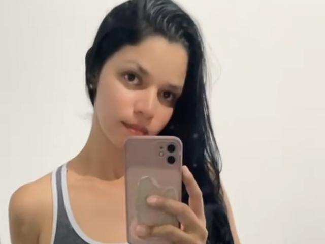 EvaYRox, con lunghi capelli neri, indossa una canotta grigia, tiene un telefono rosa davanti al viso, sta scattando un selfie allo specchio, mostrando il seno..