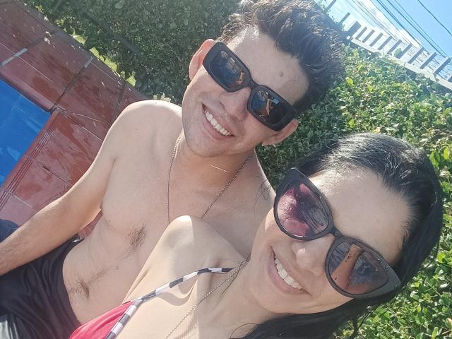EvaYRox mit dunklem Haar und Brille, lächelnd, oben ohne, zeigt kleine Brüste und einen winzigen, pinken Bikini-Unterteil, steht am Pool neben einem Mann ohne Hemd, beide tragen Sonnenbrillen..