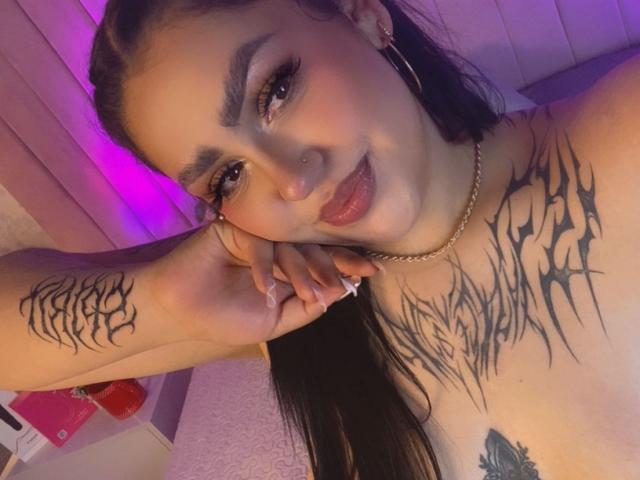 EmmaMyles with black tattoos on chest and arm, long dark hair, hoop earrings, red lipstick, and a cute smile, showing her finger with white nail. Purple lighting.