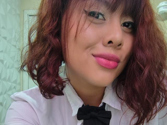 Noemi Thornee tiene el pelo rojo y ondulado, lápiz labial rosa, una pajarita negra, una camisa blanca y un piercing en la nariz, y está sonriendo con los labios ligeramente levantados. No se ven senos, glúteos ni vulva visibles..