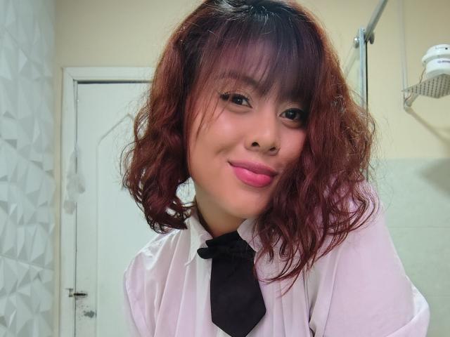 Noemi Thornee, con cabello rizado rojo, vestida con una camisa blanca y una corbata negra, lleva lápiz labial rosa y está sonriendo a la cámara. Tiene el nose perforado..