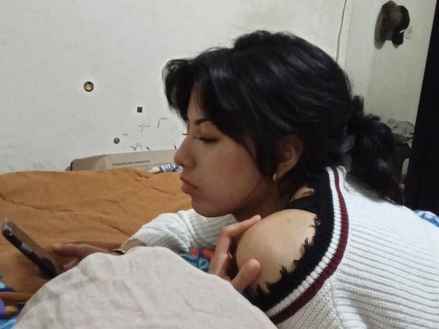 BrigidTerri with black hair, pouty face, wearing a torn white sweater, holding a phone, lying on a brown bed, looking to the side, showing her bare shoulder.