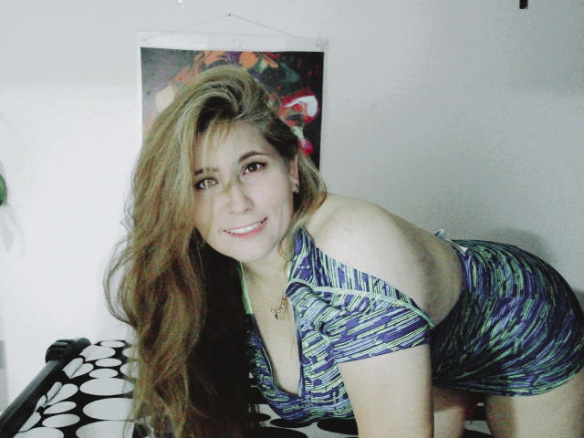 AishaSmithy, with long brown hair, leans forward, smiling, wearing a blue and green patterned dress with side cutouts, exposing her tits. She's standing in front of a wall.