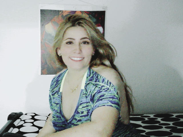 AishaSmithy with long brown hair, smiling, wearing a blue-green patterned top with a deep V-neck, sitting on a black-and-white patterned surface. Boobs partially visible.