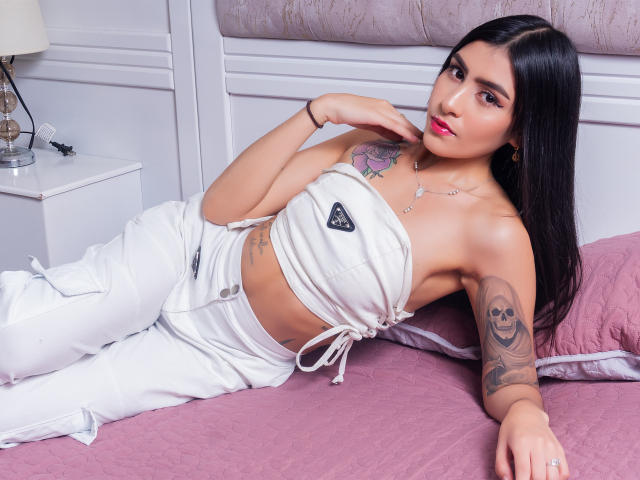 AlysonMiller with long black hair, white strapless top, white pants, tattoos on arm and chest, lying on pink bed, hand near face, pink lipstick, ring on finger.