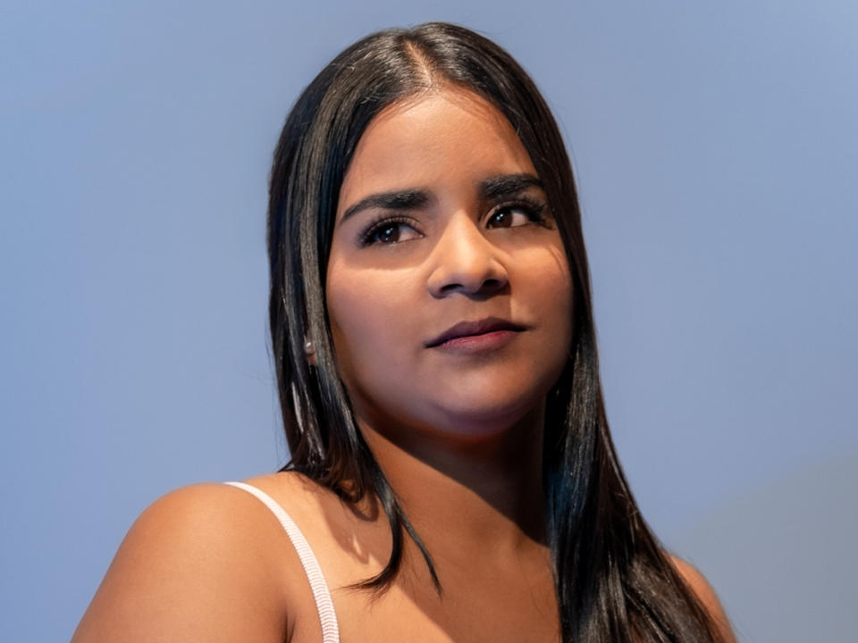 Zoe Coronado tem cabelo preto e liso, olhos castanhos e um sorriso discreto. Ela está vestindo uma blusa branca com uma alça visível. Sua pele é bronzeada e lisa..