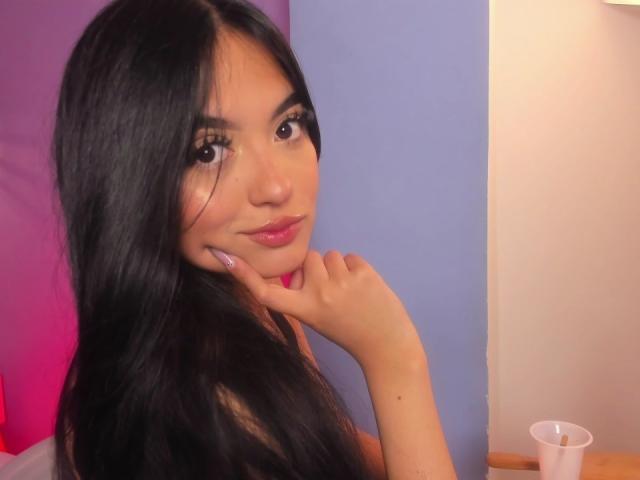 AmySaens has long, straight black hair, dark brown eyes with heavy, glittery eyeliner, and glossy pink lipstick. She's holding her chin with her right hand, which has purple nail polish. She's looking slightly to the right with a subtle, confident smile. The background has a pink and blue gradient. A white plastic cup is on a wooden surface in the lower right corner. The lighting is soft and warm.