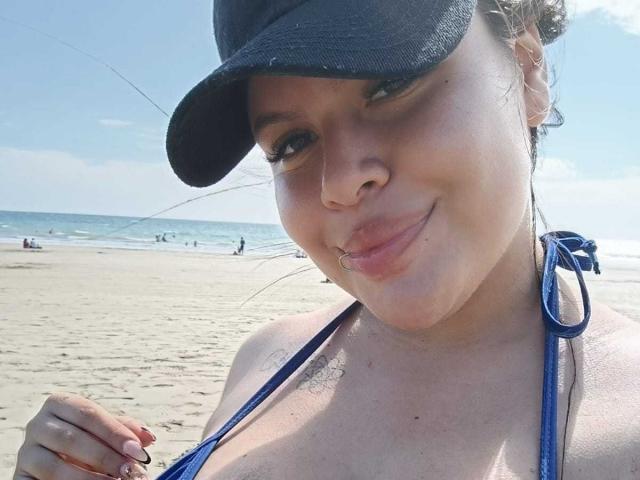 PlayfulCurvy, wearing a blue bikini top and black cap, has medium tits, a nose ring, and a subtle smile, standing on a sandy beach with the ocean in background.
