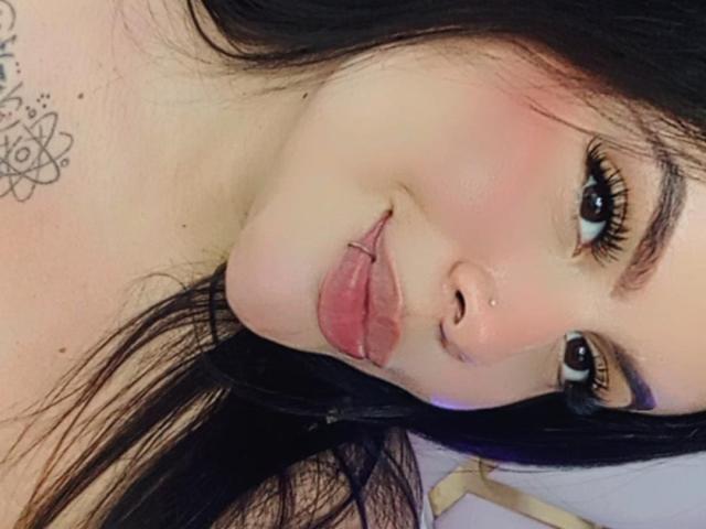 PlayfulCurvy has pouty pink lips, a septum piercing, and black eyeliner, with long black hair and a tattoo on her neck. She's looking at the camera with a slight smile.