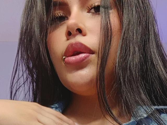 PlayfulCurvy with long brown hair, slightly parted lips, and a lip piercing, looking at the camera with one eye closed, wearing a blue shirt, hand on chest.