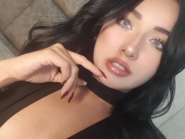 LillyTaylor with black hair, blue eyes, and glossy pink lips, touching her neck with red-manicured fingers, wearing a black top, showing her soft, fair skin.