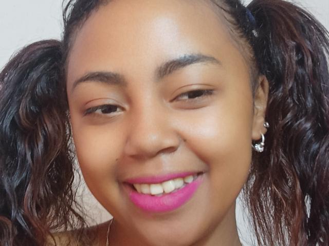 MaliciaQueen's face with brown skin, pink lipstick, curly black hair in pigtails, small nose, brown eyes, and a subtle smile, wearing a diamond earring on her right ear.