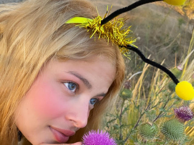 ShantalSummer with blonde hair, pink cheeks, and blue eyes, wearing a yellow and black flower headband with yellow pom-poms, looking at purple thistles.
