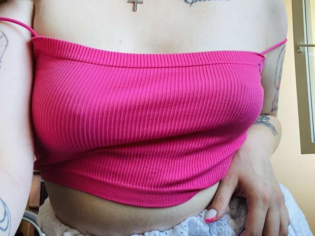 KarKutie's pink ribbed tube top showing her big, round tits with a cross tattoo above, pink strap, white lace bottom, and tattooed arms, holding her soft belly.