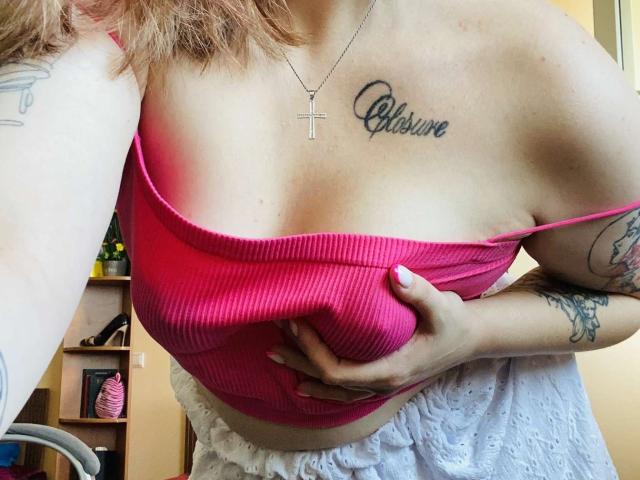 KarKutie's large tits in a pink ribbed tank top, holding one breast with a hand, tattoo of "Chasline" above her right boob, cross necklace, white lace bottom.
