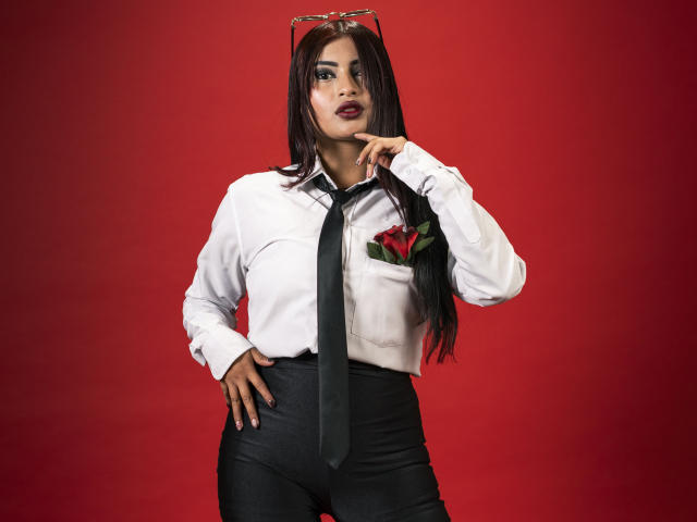 EmmaaRodriguez with long black hair, white shirt, black tie, and black pants, holding glasses on head, red rose in pocket, dark lipstick, hand on hip, red background.
