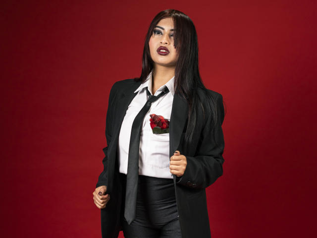 EmmaaRodriguez with long black hair, dark lipstick, black suit, white shirt, black tie, and red rose in pocket, holding suit jacket, red background. Her ass is covered by black pants.