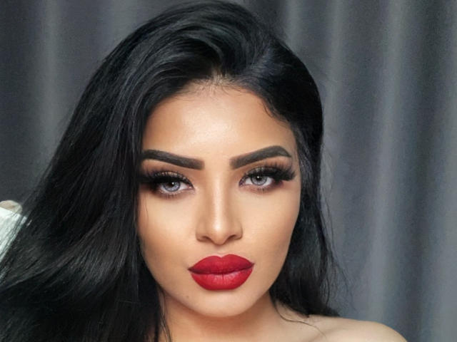Close-up of Devasha with long, straight black hair. She has bold, dark eyebrows, and grey eyes with heavy black eyeliner and long lashes. Her skin is smooth and light brown. Devasha's red lipstick is vivid and slightly matte, contrasting with her neutral-toned skin. The lighting is even, highlighting her facial features. The background is a blurred grey, keeping focus on her face. The image has a sharp, realistic style.