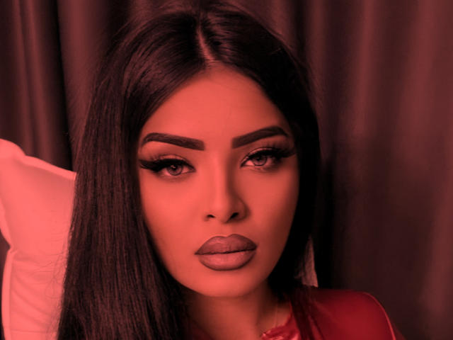 Close-up of Devasha with dark, straight hair, full eyebrows, and heavy eyeliner. Her skin is smooth, and she has glossy, matte purple lipstick. She wears a red garment. The warm, reddish light casts a soft glow on her face. Her eyes are slightly down, creating a sultry expression. The background is blurred, focusing attention on Devasha's face and makeup. The image has a high-contrast, almost neon-like quality.