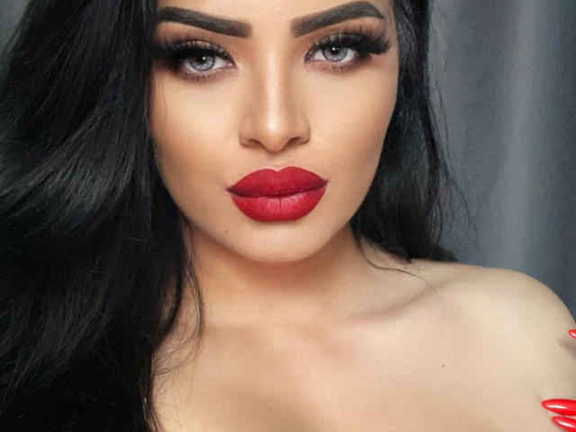 Close-up of Devasha with dark, wavy hair. She has thick, arched eyebrows, dark eyeliner, and long, thick eyelashes. Her gray eyes are slightly downcast. Devasha wears bright red lipstick that stands out against her tan skin. Her bare shoulders are visible. Her long, red-painted nails are in the bottom right corner. The image has a sharp focus on her face, highlighting her makeup and lipstick.