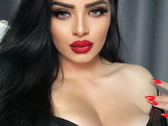 Devasha with red lipstick, dark eyebrows, long black hair, and red nails, showing off her big tits in a black top. She's staring with blue eyes, looking sexy.