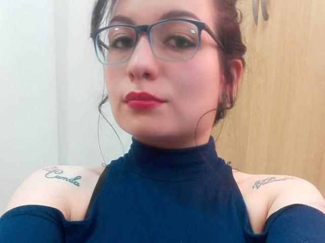 ElizabethDoom with glasses, red lipstick, and dark blue off-shoulder top, showing tattoos on her shoulders, looking at the camera with a slight smile. Her tits are partially visible.